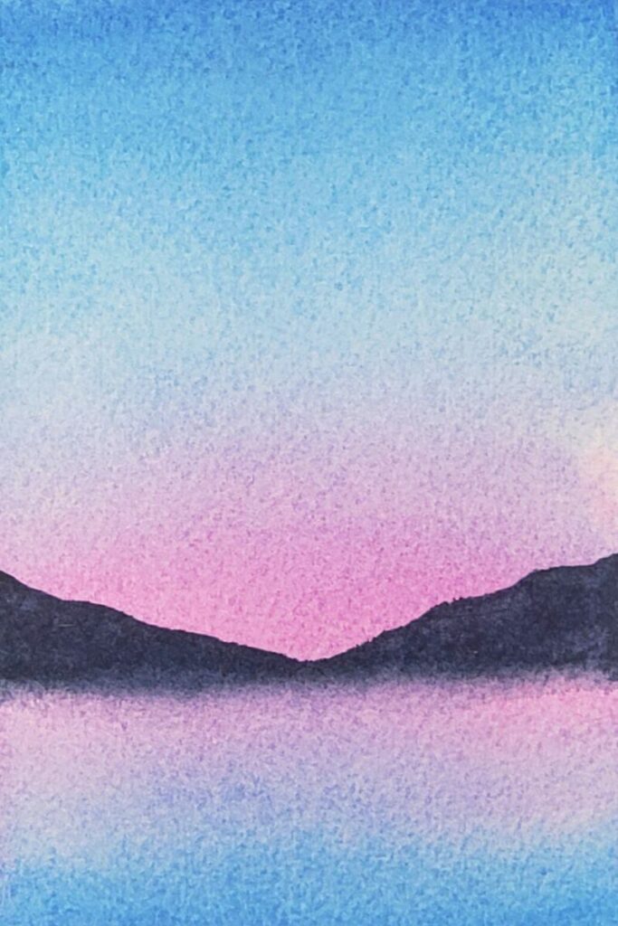 To create this easy watercolour painting, the second step is mix blue with dark grey to create indigo. Then, draw a horizon line. After, paint sloping mountains with the lowest point in the middle of the painting. Fill in the shapes with pigment and then use clear water to blend the bottom of the mountains into the pink pigment.