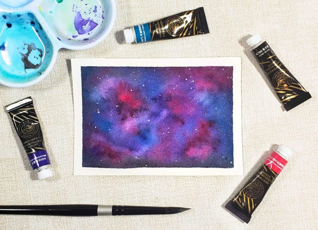 There is an image of a watercolour galaxy painting that was created by using the Paul Rubens 4th generation watercolours. It combines pink, blue, purple, and black watercolours to create the galaxy. Also, white gouache was used to create little stars.