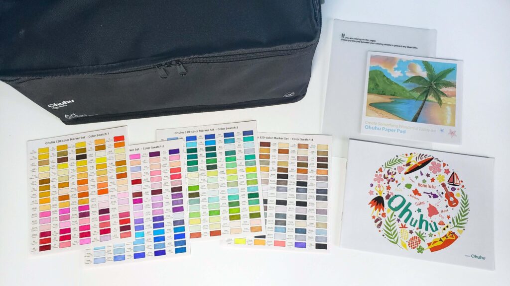 This is an Ohuhu alcohol markers review of the Honolulu set of 320 markers. In the image is a black carrying case, four colour swatch cards, a plastic sheet, a printed colour chart, and a user manual.