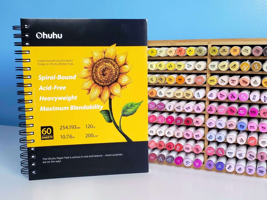There is an image of a black and yellow spiral sketchbook. Beside it is a bamboo marker organizer that's filled with yellow, orange, red, and pink Honolulu alcohol markers by the brand Ohuhu.